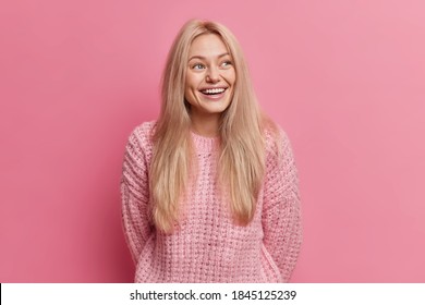 Joyful blonde woman smiles sincerely looks away with glad expression recalls something funny happened dressed in knitted jumper isolated over pink background laughs from amusement has thoughtful look