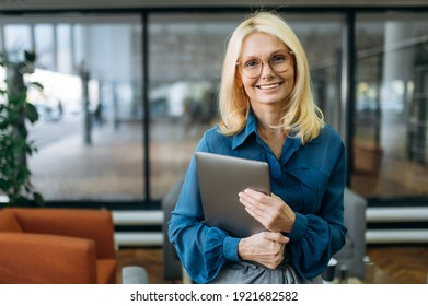 Joyful blonde-hair business woman in eyeglasses is looking at the camera, holding laptop in arms and smiling. Beautiful middle aged lady in stylish wear stands in modern office - Powered by Shutterstock