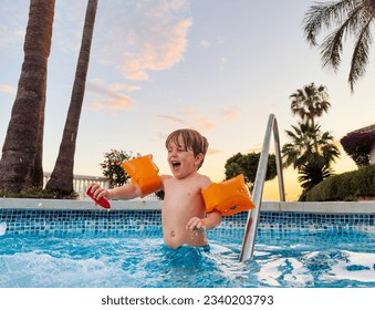 Joyful boy in arm bands enjoying sunset swimming pool having fun laughing and splashing