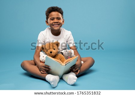 Joyful boy of elementary age looking at you with toothy smile while sitting on the floor in studio, reading book and playing with soft toys