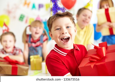 Joyful boy with giftbox looking at camera with his friends on background