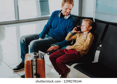 Joyful Boy Is Sitting At Airport And Waiting For Flight With Father. He Is Speaking On Cell Phone. Parent Is Watching Him While Using Headphones