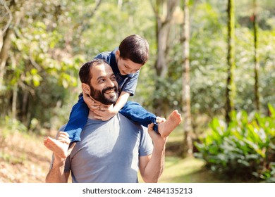 A joyful Brazilian father carrying his young son on his back in a lush, green forest. They are laughing and playing, highlighting a special moment of father-son bonding on Father's Day - Powered by Shutterstock