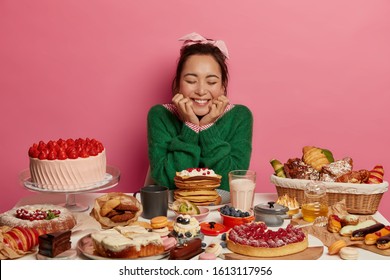 Joyful brunette Asian woman in green jumper dreams about eating tasty desserts, being sweet tooth, has sweet life, models over rosy wall, has yummy breakfast. Unhealthy eating and junk food concept