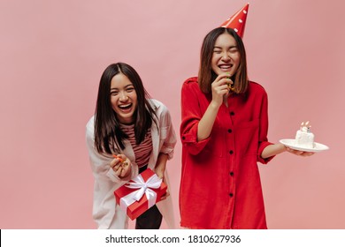 Joyful Brunette Asian Women Laugh On Pink Isolated Background. Lady In Party Hat And Red Blouse Holds Birthday Cake. Girl In Striped Shirt Poses With Gift Box.