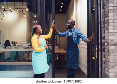 Joyful business partners in uniform looking at each other and giving high five while standing on entrance of own cafe and celebrating successful start