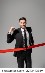 joyful businessman looking at camera and showing triumph gesture near red finish ribbon isolated on grey - Shutterstock ID 2293443989
