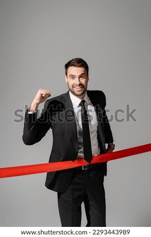 joyful businessman looking at camera and showing triumph gesture near red finish ribbon isolated on grey