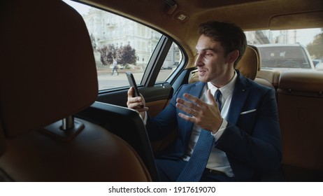 Joyful businessman telling good news in video message on smartphone in business car. Happy male professional recording motivating video in modern car. Cheerful man having video chat in automobile.