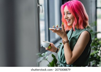 joyful businesswoman with pink hair and piercing holding cup of coffee while recording voice message on smartphone