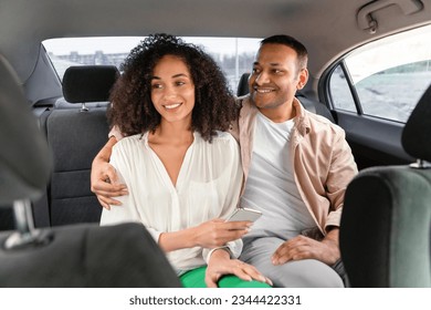Joyful Cab Experience. Smiling Arab Passengers Couple Enjoying Comfortable Car Ride In Taxi, Lady Holds Smartphone with Transfer Transportation Application, Spouses Hugging On Backseat In Auto