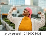 Joyful cancer survivor in her 30s, wearing a pink headscarf and yellow sweater, flexing her muscles in an urban setting.