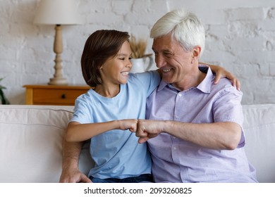 Joyful Carefree Small Preteen Kid Boy Bumping Fists With Caring Loving Older Mature Retired Grandfather, Coming To Common Decision, Making Deal Or Celebrating Good News, Resting On Couch At Home.