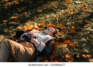 Joyful Carefree Young Woman Hipster Photographer Lying On Fallen Colorful Foliage Outdoors. Laughing Woman Tourist With Vintage Camera Relaxing Enjoying Autumn Day. Travel, Hobby, Lifestyle Concept.