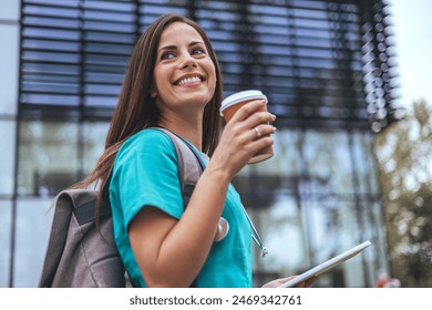 A joyful Caucasian female nurse enjoys a break outside a medical facility, holding a digital tablet in one hand and a coffee cup in the other, dressed in scrubs with a stethoscope around her neck. - Powered by Shutterstock