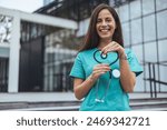 A joyful Caucasian female nurse forms a heart with her stethoscope, displaying care outside a medical facility, dressed in scrubs, symbolizing compassion in healthcare.