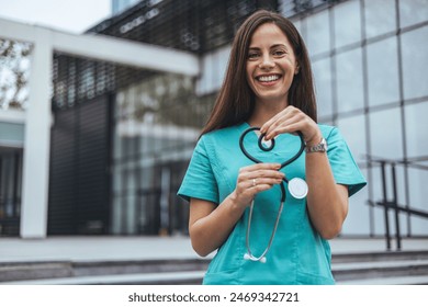 A joyful Caucasian female nurse forms a heart with her stethoscope, displaying care outside a medical facility, dressed in scrubs, symbolizing compassion in healthcare.