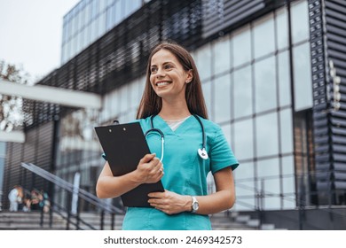 A joyful Caucasian female nurse stands confidently outside a hospital, holding a clipboard while dressed in teal scrubs with a stethoscope around her neck - Powered by Shutterstock