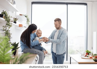 Joyful caucasian man and woman making funny layers out of palms with small child at home. Loving couple entertaining cute baby girl with hand stacking game while waiting for dinner in kitchen.