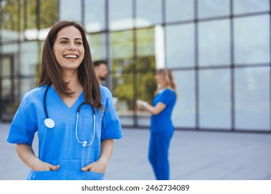 A joyful Caucasian nurse in blue scrubs stands confidently outside a hospital, stethoscope around her neck, with her colleagues in the background. - Powered by Shutterstock