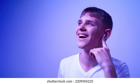 Joyful Caucasian Teenage Boy Pushing On Wireless Earphone For Choosing Music. Blonde Guy Of Zoomer Generation. Modern Youngster Lifestyle. Blue With White Light Background. Studio Shoot. Copy Space