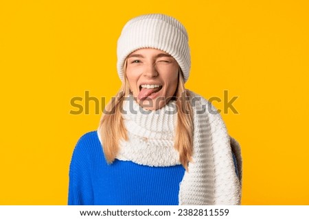 Joyful caucasian woman in blue sweater playfully sticks out her tongue and winking while wearing white scarf and hat, posing vibrant yellow background
