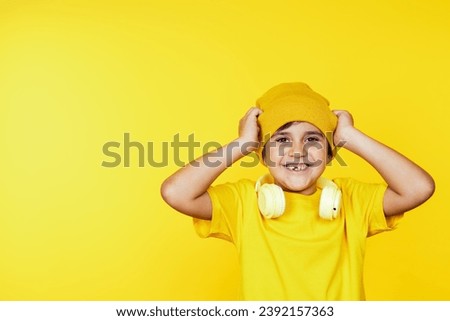 Joyful child in yellow attire adjusting beanie, white headphones around neck against a bright backdrop.