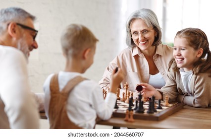 Joyful children brother and sister playing chess while sitting in living room with senior grandparents while spending time together on weekend, kids sitting at table with chessboard and smiling