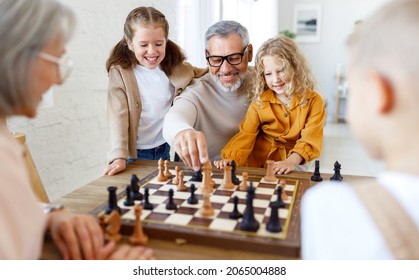 Joyful children brother and sister playing chess while sitting in living room with senior grandparents while spending time together on weekend, kids sitting at table with chessboard and smiling - Powered by Shutterstock