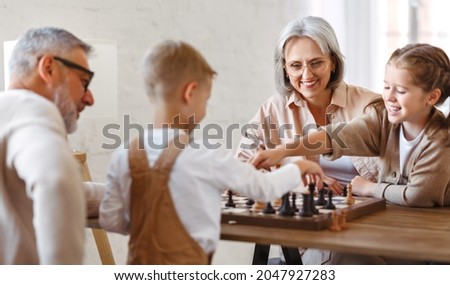 Joyful children brother and sister playing chess while sitting in living room with senior grandparents while spending time together on weekend, kids sitting at table with chessboard and smiling