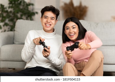 Joyful Chinese Couple Playing Video Game Holding Joysticks Sitting On Sofa At Home On Weekend. Asian Boyfriend And Girlfriend Play Videogames. Family Fun, Computer Gaming And Games Concept