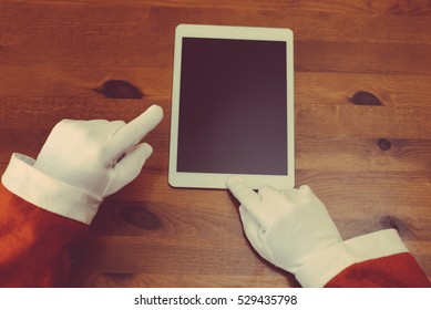 Joyful Christmas online shopping. Closeup on Santa Claus working using mobile tablet computer on wooden table background. Social network, electronic commerce, banking, logistic operations, resolution