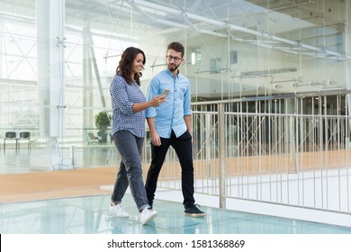 Joyful colleagues in casual looking at cellphone screen together while going through office hallway. Young man and woman walking indoors with glass wall in background. Communication concept - Powered by Shutterstock