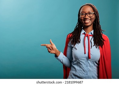 Joyful And Confident Superhero Woman Wearing Red Cape While Pointing Fingers To Left On Blue Background. Selfless And Strong Young Adult Justice Defender Person Posing At Camera. Studio Shot.