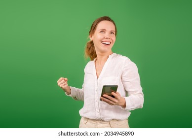 Joyful, Contented Woman Of Middle Age 40 Years Old In A White Shirt With A Mobile Phone On A Green Background. Online Shopping, Happy News Concept.