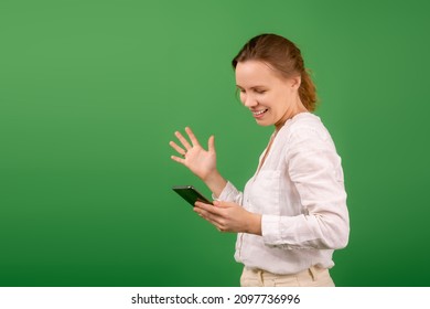 Joyful, Contented Woman Of Middle Age 40 Years Old In A White Shirt With A Mobile Phone On A Green Background. Online Shopping, Happy News Concept.