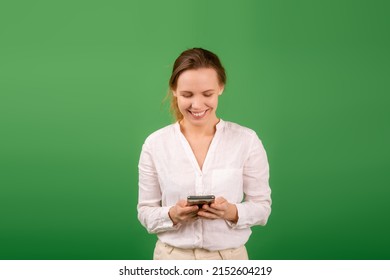 Joyful, Contented Woman Of Middle Age 40 Years Old In A White Shirt With A Mobile Phone On A Green Background. Online Shopping, Happy News Concept.