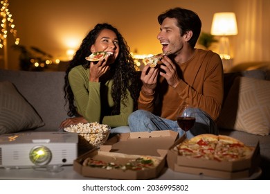 Joyful Couple Eating Pizza And Laughing Watching Comedy Film Using Home Cinema Projector Sitting On Couch Indoor. Entertainment And Fun Concept. Family Enjoying Movie On Weekend