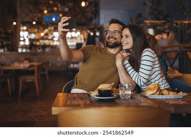 A joyful couple embraces while taking a selfie in a warmly lit restaurant. Plates of food surround them, creating a cozy and relaxed atmosphere. Perfect for concepts of love, dining, and togetherness. - Powered by Shutterstock