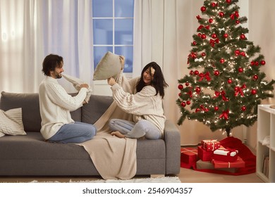 A joyful couple engages in a playful pillow fight on a sofa next to a decorated Christmas tree. Perfect for holiday ads, showcasing fun, laughter, and festive spirit in a cozy setting. - Powered by Shutterstock