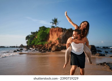 Joyful couple enjoys piggyback ride on tropical beach, woman raises hand high, man carries partner by sea. Romantic getaway, playful mood, sandy shore, love adventure. Tourists explore island paradise - Powered by Shutterstock