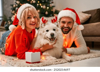 A joyful couple in festive Santa hats poses with their fluffy dog in reindeer antlers, surrounded by a beautifully decorated indoor space, enhancing the cheerful holiday atmosphere - Powered by Shutterstock