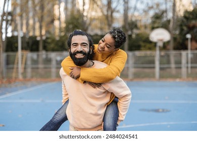 Joyful couple in their 30s enjoying a playful piggyback ride on an outdoor basketball court, smiling and laughing. - Powered by Shutterstock