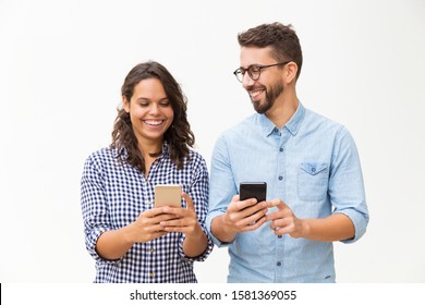 Joyful Couple Using Smartphones, Talking And Laughing. Young Woman In Casual And Man In Glasses Posing Isolated Over White Background. Wireless Internet Concept