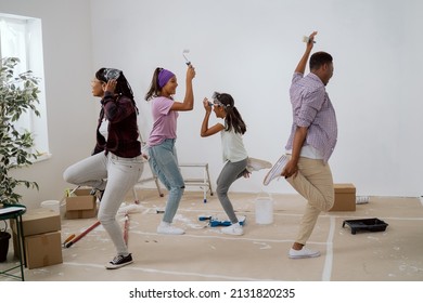 Joyful crazy family painting the walls with white paint with brushes, they are dancing in the middle of the room, singing, parents are playing with their children, the house prepared for renovation