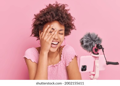 Joyful curly Afro American woman influencer laughs happily keeps hand on face has funny conversation with subscribers poses against pink backgroud. Socal media blogging and technology concept - Shutterstock ID 2002155611