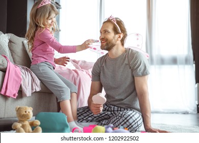 Joyful dad is playing with his little daughter in fairy. Girl is painting his lips by cosmetic and smiling. Man is sitting on floor and wearing fairy wings and crown  - Powered by Shutterstock