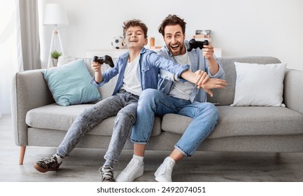 Joyful dad and son with joysticks playing video games at home, boy distracting father with hand, faving fun together, sitting on sofa in living room. Fatherhood and leisure activities concept - Powered by Shutterstock