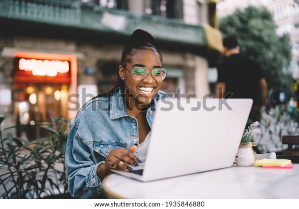 Joyful dark skinned female blogger typing text of\
publication using laptop computer working remotely, cheerful\
African American woman in spectacles share positive content and\
media files on netbook