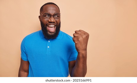 Joyful dark skinned man does winner gesture celebrates victory makes fst bump exclaims from happiness wears round spectacles basic blue t shirt isolated over beige background blank copy space